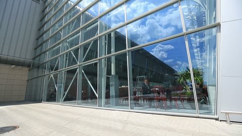 Young business man waits for plane at airport, sitting in bright cafe. Blue clouded sky in reflection of mirroring outer glass wall