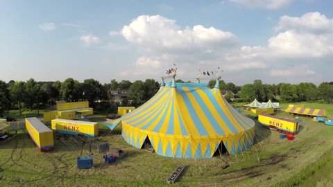 Aerial circling around circus tent colored in blue and yellow stripes also showing circus trucks and smaller tents and buildings on top of structure some flags are placed moved by wind 4k resolution