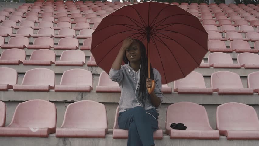 Sad african american young woman in uniform with umbrella on the stadium