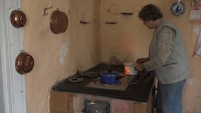 elderly countrywoman put rustic pot on old rural clay stove in kitchen. 4K UHD video clip.