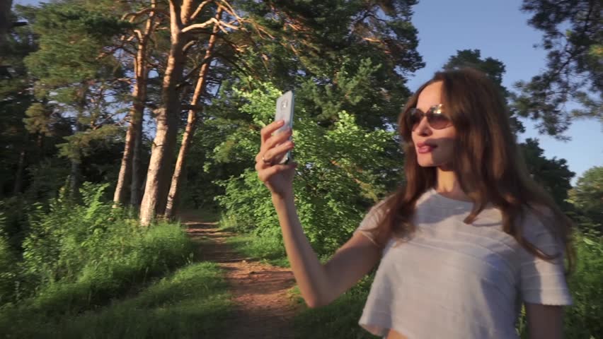 Young long hair brunette girl in sunglasses making selfie with her phone in sunset forest