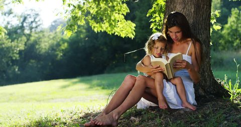 a happy young mother with the child under a tree in the fresh shade reads a fairy tale to her little girl. concept of childhood. happy children and youth and happiness. nature shoot on red camera 