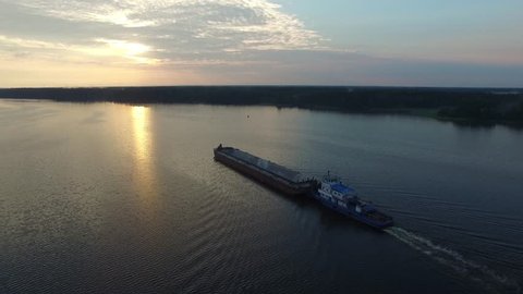 Cargo transport ship sailing on a wide river. Summer dawn. The camera moves through the air away from the ship. The island in the middle of the river. Sun beautifully reflected in the water.