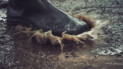 Black rubber boots step on a puddle and creates a splash which flies around. Slow motion, high speed camera, 250fps