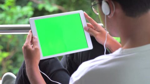 man Sits on chair Using Tablet PC Smart Phone Mobile Device in Nature with Green Screen and wearing headphones.