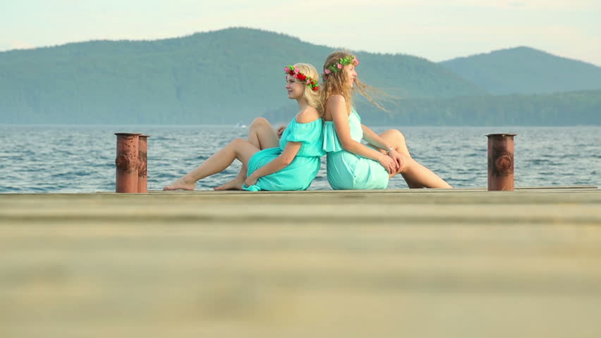 Cute girls with wreaths on their heads are resting on the river. Two young beautiful woman in a blue dress sits on a wooden pier on the river.