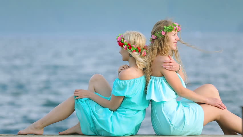 Cute girls with wreaths on their heads are resting on the river. Two young beautiful woman in a blue dress sits on a wooden pier on the river.