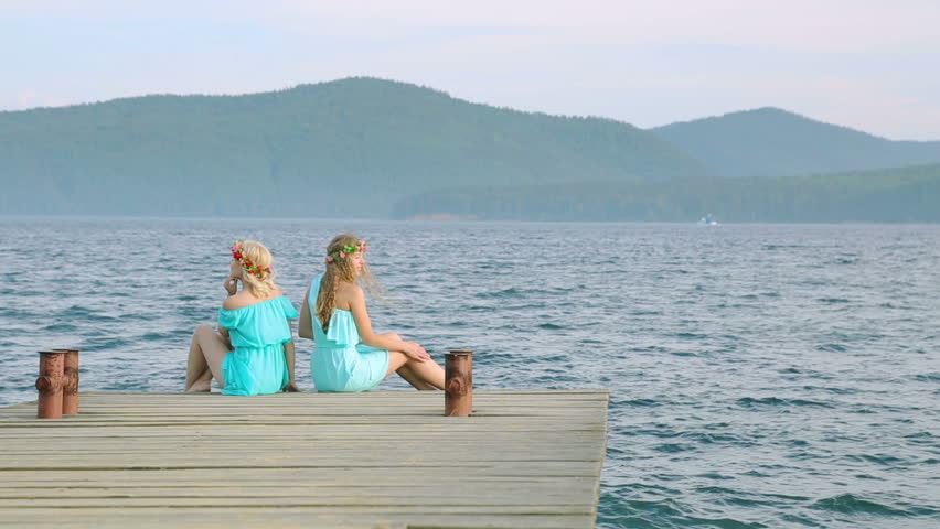 Cute girls with wreaths on their heads are resting on the river. Two young beautiful woman in a blue dress sits on a wooden pier on the river.