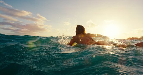Surfer paddling over blue ocean wave at sunset in slow motion, outdoor fitness lifestyle