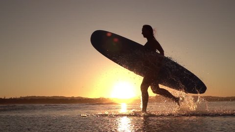 SLOW MOTION SILHOUETTE: Young surfer girl enjoying seaside summer vacation activities, holding longboard surfboard and running in shallow water in beautiful ocean at amazing golden light sunset