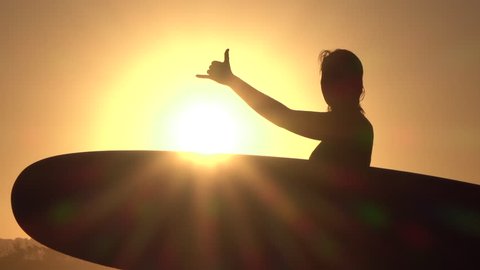 SLOW MOTION CLOSE UP SILHOUETTE: Cheerful happy young surfer girl holding longboard surfboard and doing the shaka surf culture sign gesture with fingers at golden sunset in surfing town Byron bay