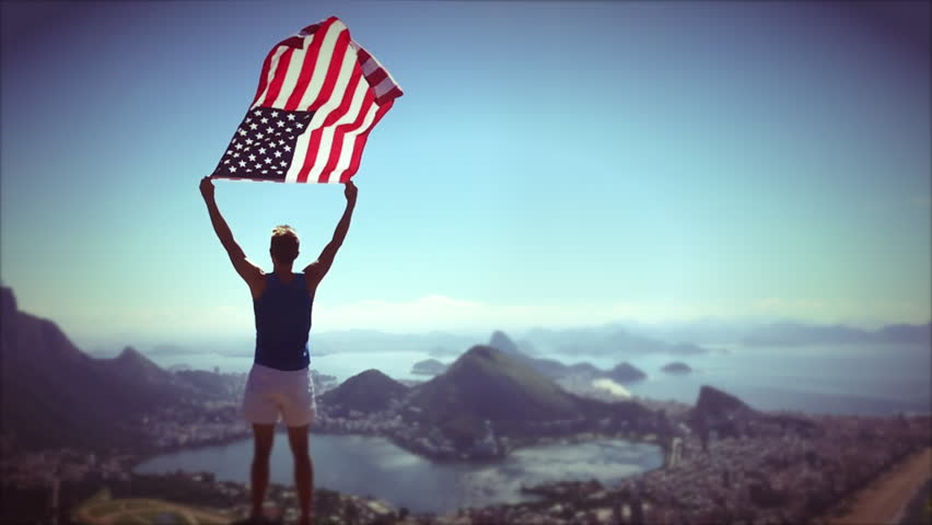 Athlete stands holding an American flag waving in slow motion at a bright overlook of the city skyline of Rio de Janeiro, Brazil