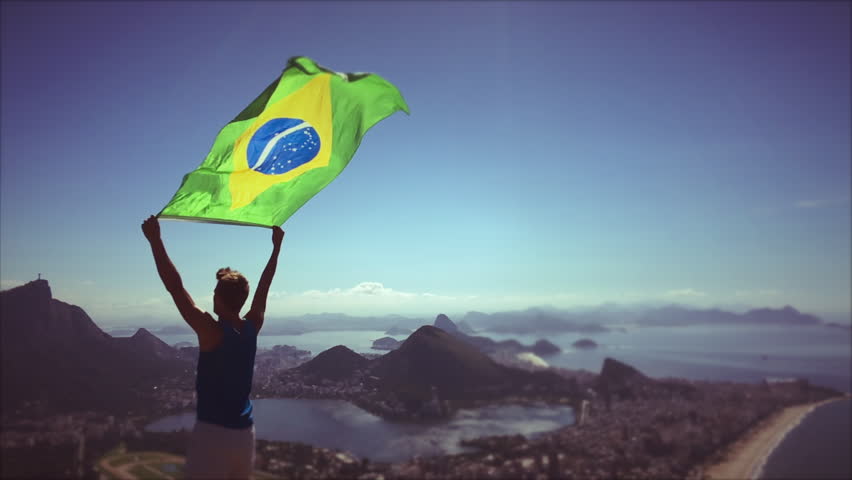 Athlete stands holding a Brazilian flag waving in slow motion at a bright overlook of the city skyline of Rio de Janeiro, Brazil Royalty-Free Stock Footage #18005965