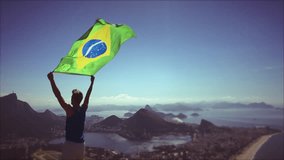 Athlete stands holding a Brazilian flag waving in slow motion at a bright overlook of the city skyline of Rio de Janeiro, Brazil