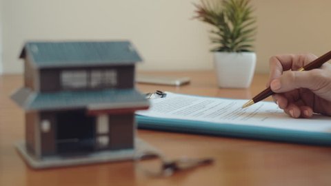 Female real estate agent reading and signing ownership contract sitting at the desk with house model and key. Concept of property loan and finance. 