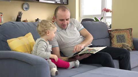Cute toddler girl climb on sofa, sit near dad and read book with images together. Child and man showing book with finger. Static closeup shot.
