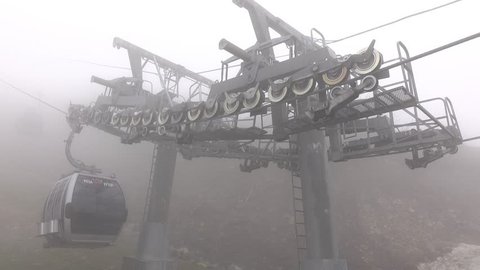 MDG cabin pass by support pylon, bracket ride over many steel wheels. Mechanism hold haul steel cable. Cloudy air at high altitude, misty appearance. Close shot, camera look to sheave system atop