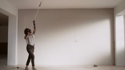 Funny curly man wears pants with suspenders acts like he is dancing waltz while painting celling with brush roller and white paint Home diy renovation
