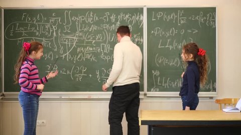 Two girls and man talking and writing on blackboard with equations.