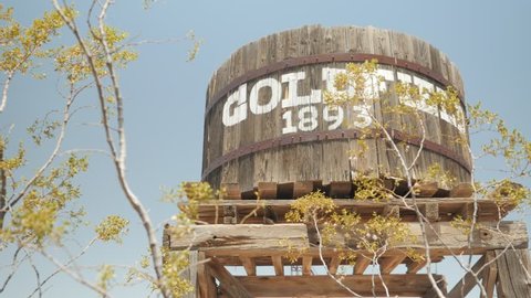 Goldfield Ghost Town, AZ, USA - July 16, 2016: Goldfield 1893 Ghost Town sign on a water barrel behind tree branches and leaves.