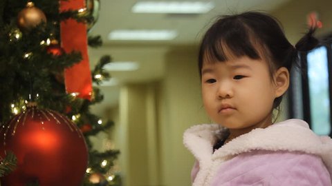 Beautiful Baby Asian Toddler Girl in Purple Coat Looking at Christmas Ornaments on Tree