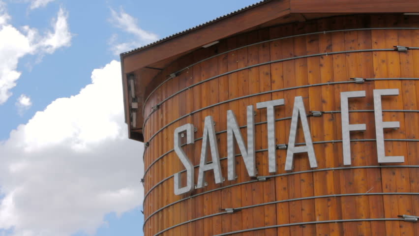 Establishing shot of a rustic wooden water tower in Santa Fe, New Mexico. Puffy white clouds float in the blue sky.
