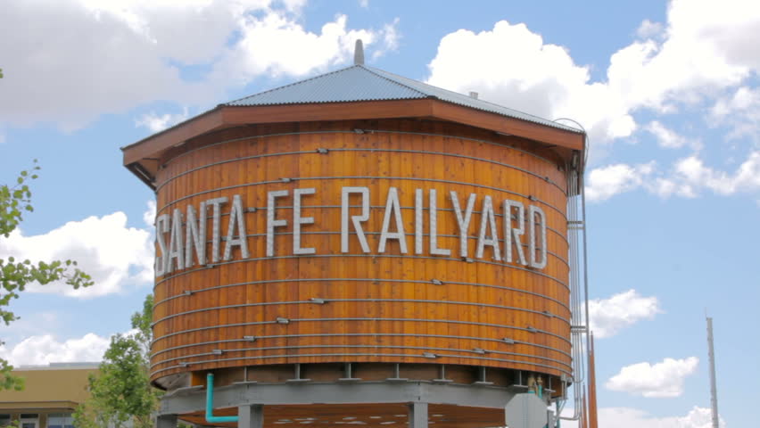 Wide shot establishes the Santa Fe Rail Yard area in New Mexico. The large rustic wooden water tower has a sign that reads Santa Fe Railyard. Fluffy white clouds float lazily in the blue summer sky.