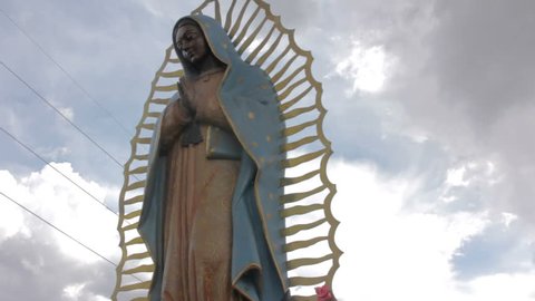 Low angle shot of of a statue of the Virgin of Guadalupe giving the symbol an imposing feeling. Dark clouds gather ominously behind the beautiful Mexican Catholic religious icon.