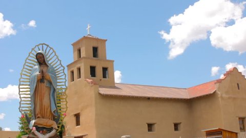 Extreme wide shot of a statue of Our Lady of Guadalupe the Patron Saint of Mexico. The religious icon stands peacefully in front of a historic adobe church in Santa Fe, New Mexico.