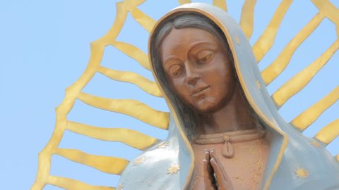Closeup of of a statue of the Virgin of Guadalupe. The low angle of the religious icon symbolizes peace and serenity. A cloudless blue sky sits behind the beautiful Mexican Catholic saint.