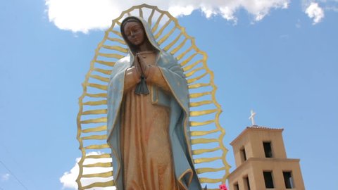 Low angle shot of a statue of the virgin of Guadalupe standing serenely in front of an adobe church. Blue sky with clouds is in the background behind the religious icon and the adobe Catholic church.
