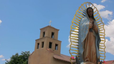 Panning shot of a statue of Our Lady of Guadalupe the Patron Saint of Mexico. The religious icon stands peacefully in front of a historic adobe church in Santa Fe, New Mexico. 