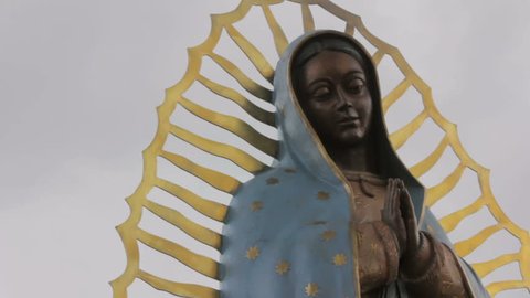 Closeup of of a statue of the Virgin of Guadalupe. The low angle of the religious icon symbolizes an imposing feeling. Dark clouds gather ominously behind the beautiful Mexican Catholic saint.
