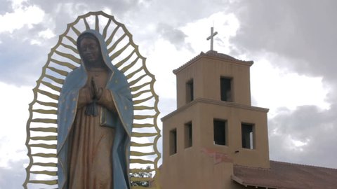 Wide shot of a statue of the virgin of Guadalupe standing serenely in front of an adobe church. Ominous storm clouds gather behind the religious icon and the Mexican Catholic church.