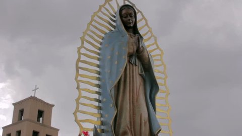 Wide shot of a statue of the virgin of Guadalupe standing in front of an adobe church. Low angle of the catholic saint has a foreboding feeling. Ominous storm clouds gather behind the religious icon.