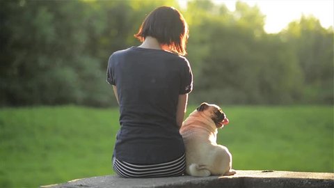 Happy young woman sit back with dog and looking at each other outdoors, girl stroking her mops in a park, warm sunny day