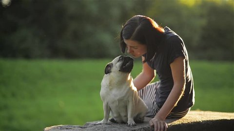 Young beautiful woman blowing a kiss to her dog at the park, Cute pug kissing a girl, dog and owner sitting on the parapet