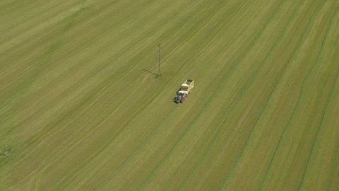 AERIAL, DISTANCING: Flying above tractors picking hay on big vast grass field and collecting haystack in forage wagon. Farmer working on big agricultural farmland near suburb district outside town