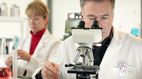 A woman and man who appear to be scientists or chemists working in a laboratory using microscopes and other technologies.
