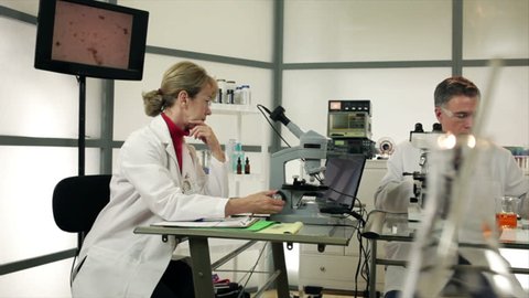 A male lab tech using a Pipette and a female researcher with a microscope camera working together in a laboratory setting. Slow dolly movement past colorful objects in the foreground.