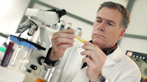A handsome mature scientist or chemist sitting in a laboratory visually examining a liquid he has in a test tube.