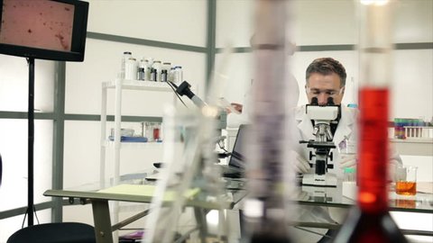 A male researcher using a microscope and a female scientist who comes from the back of the lab then sits and continues working. Slow dolly movement past colorful objects in the foreground.