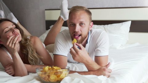 The girl and the guy eating chips lying on the bed