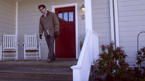A sad man on the verge of tears leaves his modern, contemporary, generic, house with a red door looking disheveled and frustrated
