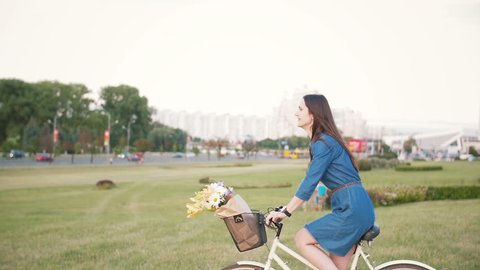 Side view of a brunette girl with long hair riding a bike in the city with flowers in a basket, slow mo, steadicam shot