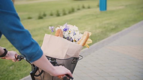 Girl hands on a handlebar of a bicycle, flowers and bread in a basket, cycling in the city, slow mo, steadicam shot