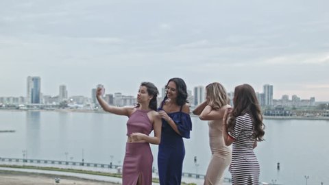 Four Attractive young women in party dresses on high hill get a selfie, wide shot