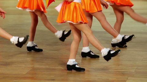 Five girls in shoes with taps and orange skirts tap dance, only legs are visible