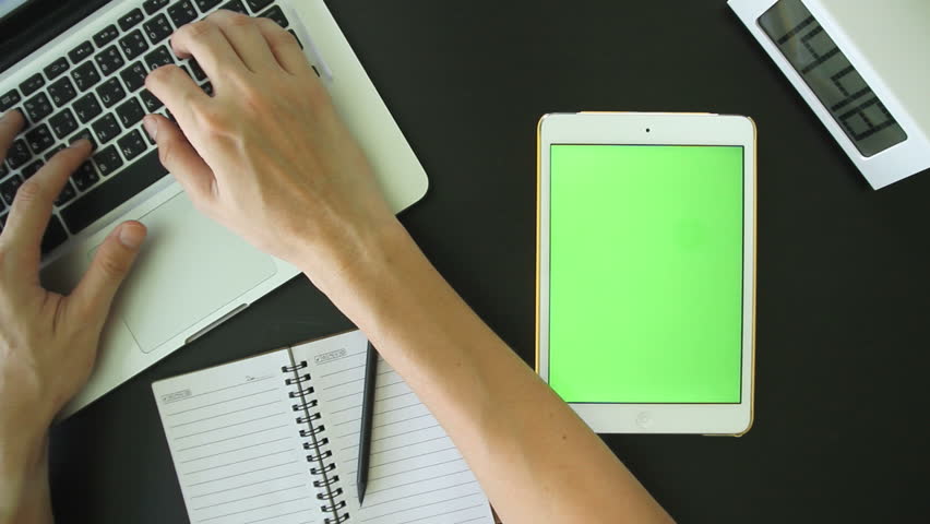 Top view of man working on laptop and white tablet with touch blank green screen on table.