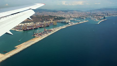 Landing in Barcelona, Spain, Catalonia. Traveling by air. Bird's-eye view through an airplane window. Flying aerial over the Barcelona city port, houses and streets. A wing is seen in left top corner.